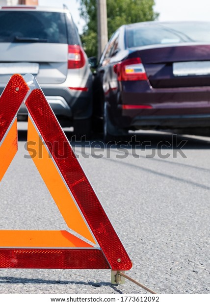 Triangular red retro-reflective sign of accident\
on the road. Collision of two cars. Broken bumper and hood. Car\
accident on the\
street