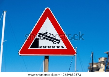 A triangular road sign with a red border warns of a nearby quay, depicting a car potentially falling into water, under clear blue sky.