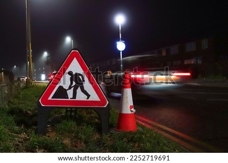 A triangular roadworks sign and traffic cone at the side of a road at night to worn motorists of roadworks ahead,