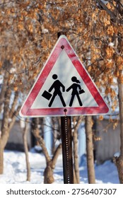 A triangular school or pedestrian crossing warning sign, featuring cracked paint, stands prominently in a snowy winter landscape.