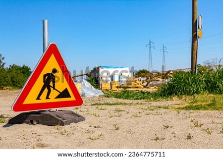 Triangular traffic sign with caution symbol of warning, work in progress, placed in front of construction site zone area.