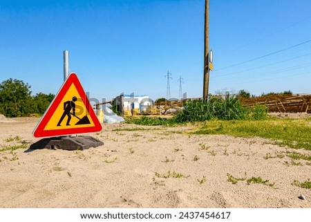 Triangular traffic sign with caution symbol of warning, work in progress, placed in front of construction site zone area.