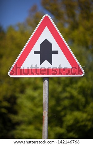 Triangular traffic sign indicating a crossroads on a main thoroughfare mounted on a roadside pole against greenery