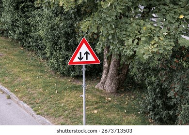 A triangular traffic sign is placed alongside a roadway, signaling a one-way traffic direction. The surrounding area features lush green grass and trees.