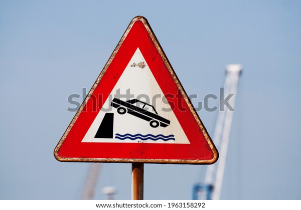 Triangular warning road sign, white with red\
outline depicting a car falling off a harbor dock due to the\
absence of protective\
barriers