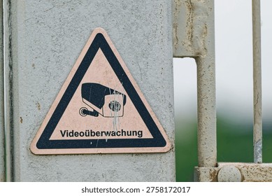 A triangular warning sign indicating video surveillance is attached to a weathered metal gate with a blurred natural background.