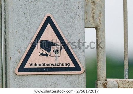 A triangular warning sign indicating video surveillance is attached to a weathered metal gate with a blurred natural background.