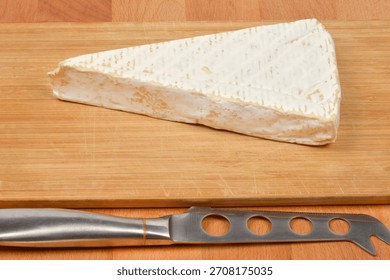 A triangular wedge of soft-ripened Brie cheese with a white rind sits on a bamboo cutting board, with a specialized cheese knife resting in the foreground