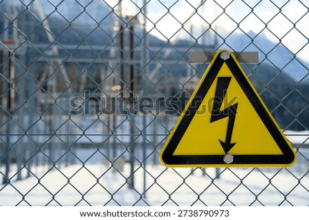 Triangular yellow high-voltage danger sign on fence, warning for electric shock, power transformer station out of focus behind, electrical safety symbol, utility infrastructure