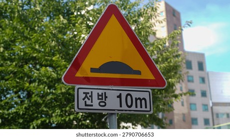 A triangular yellow and red speed bump warning traffic sign with an additional rectangular plate indicating ten meters ahead.