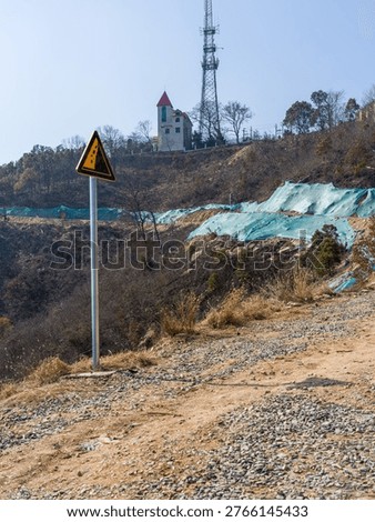 A triangular yellow warning sign stands beside a rugged dirt path on a hillside, with a building, telecom tower, and green protective netting in the background. The scene represents construction safety, rural development, and environmental modification.