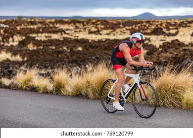 Triathlon Professional Cyclist Man Cycling Road Racing Bike In Time Trial Helmet And Compression Tri Suit In Hawaii Landscape For Kailua-Kona Ironman. Triathlete Biking In Nature.