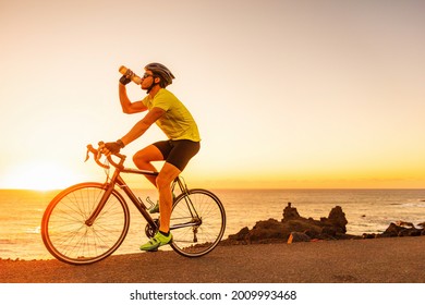 Triathlon race biking cycling sports athlete cyclist man drinking water bottle while riding road bike at sunset after training wearing cycle accessories, smartwatch, helmet, sunglasses, gloves, shoes. - Powered by Shutterstock