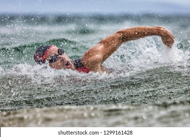 Triathlon Swim Tired Swimmer Swimming In Ocean In Wave And Rain Storm . Professional Male Triathlon Swimmer Wearing Cap, Goggles And Red Triathlon Tri Suit Training For Ironman Breathing Out Of Water.