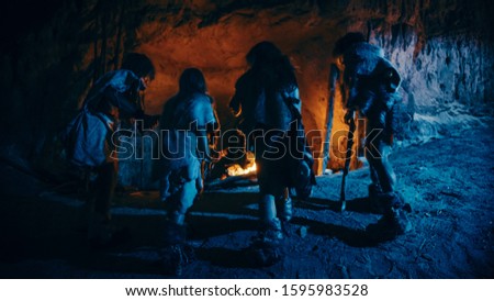 Tribe of Prehistoric Hunter-Gatherers Wearing Animal Skins Live in a Cave at Night. Neanderthal or Homo Sapiens Family Trying to Get Warm at the Bonfire, Holding Hands over Fire. Back View