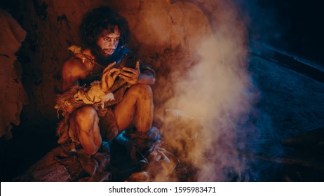Tribe of Prehistoric, Primitive Hunter Gatherer Wearing Animal Skin Uses Smartphone in a Cave at Night. Neanderthal / Homo Sapiens Male Browsing Internet on Mobile Phone, Watches Videos