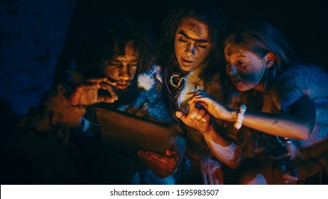 Tribe of Prehistoric, Primitive Hunter-Gatherers Wearing Animal Skins Use Digital Tablet Computer in a Cave at Night. Neanderthal or Homo Sapiens Family Browsing Internet, Watching Videos, TV Shows