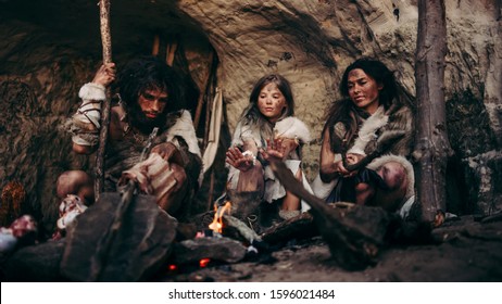 Tribe of Prehistoric PrimitiveHunter-Gatherers Wearing Animal Skins Live in a Cave at Night. Neanderthal or Homo Sapiens Family Trying to Get Warm at the Bonfire, Holding Hands over Fire, Cooking Food