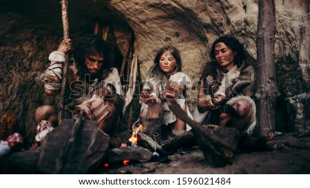Tribe of Prehistoric PrimitiveHunter-Gatherers Wearing Animal Skins Live in a Cave at Night. Neanderthal or Homo Sapiens Family Trying to Get Warm at the Bonfire, Holding Hands over Fire, Cooking Food