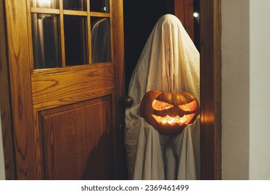 Trick or treat! Spooky ghost holding glowing Jack o lantern pumpkin in doors in dark. Scary atmospheric halloween time, person dressed as ghost trick or treating. Happy Halloween