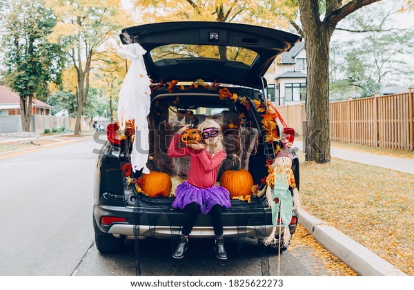 Trick or trunk. Child girl celebrating\
Halloween in trunk of car. Kid with red carved pumpkin celebrating\
traditional October holiday outdoor. Social distance during\
coronavirus covid-19.