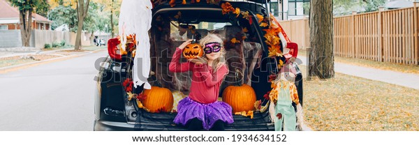 Trick or trunk. Child girl celebrating Halloween\
in trunk of car. Kid with red carved pumpkin celebrating\
traditional October holiday outdoors. Safe alternative celebration.\
Web banner header.