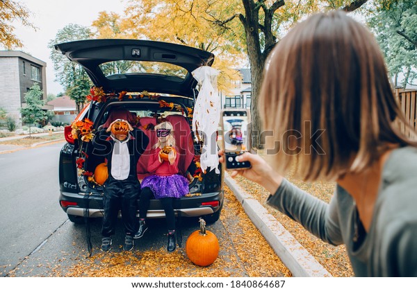 Trick or trunk. Children boy and girl with red pumpkins
celebrating October Halloween holiday in trunk of car outdoor.
Mother taking pictures of kids on smartphone camera for social
media. 