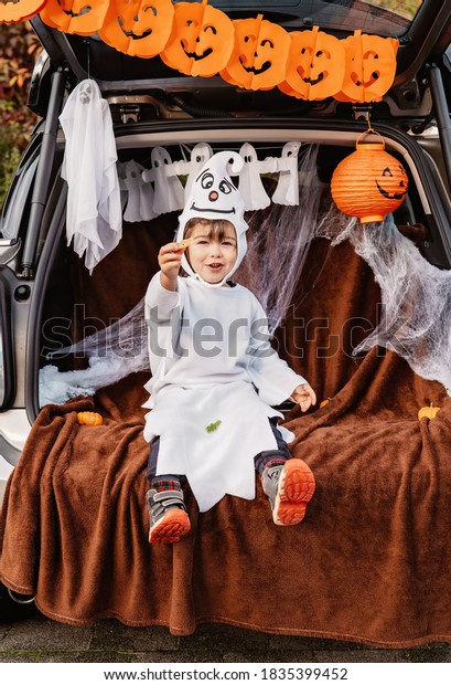 Trick or trunk. Trunk or treat. Happy child in\
ghost costume celebrating Halloween party in decorated trunk of\
car. New trend and alternative safe outdoor celebration of\
traditional holiday.