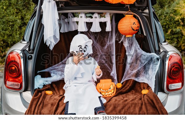 Trick or trunk. Trunk or treat. Happy child in\
ghost costume having fun celebrating Halloween party in decorated\
trunk of car. New trend and alternative safe outdoor celebration of\
traditional holiday