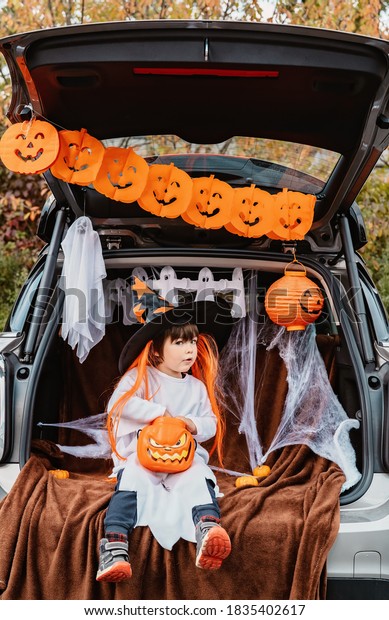 Trick or trunk. Trunk or treat. Little child in\
witch hat with funny face eating Halloween candies in decorated\
trunk of car. New trend and alternative safe outdoor celebration of\
traditional holiday