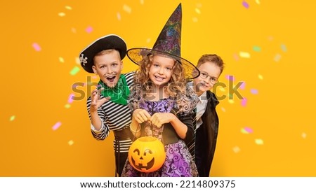 Trick-or-treating on Halloween. Three cheerful cute kids in Halloween costumes with a pumpkin basket emotionally pose trying to scare. Yellow background with colourful cofetti.