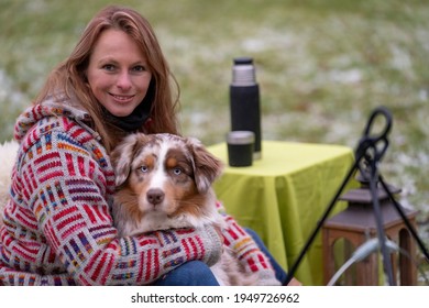 Tricolor Australian Shepherd Sits Next To A Young Woman By The Campfire. In Winter, Snow On The Grass. Cuddled With The Dog.The Fire Is Burning Under The Cauldron. Selective Focus, Thermos Coffee On