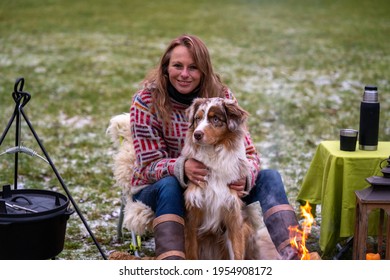 Tricolor Australian Shepherd Sits Next To A Young Woman By The Campfire. In Winter, Snow On The Grass. Cuddled With The Dog.The Fire Is Burning Under The Cauldron. Selective Focus, Thermos Coffee On