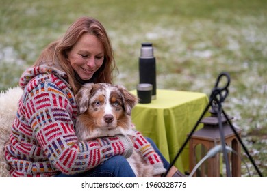 Tricolor Australian Shepherd Sits Next To A Young Woman By The Campfire. In Winter, Snow On The Grass. Cuddled With The Dog.The Fire Is Burning Under The Cauldron. Selective Focus, Thermos Coffee On