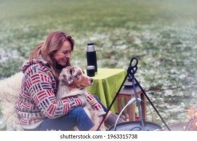 Tricolor Australian Shepherd Sits Next To A Young Woman By The Campfire. In Winter, Snow On The Grass. Cuddled With The Dog.The Fire Is Burning Under The Cauldron. Selective Focus, Thermos Coffee On