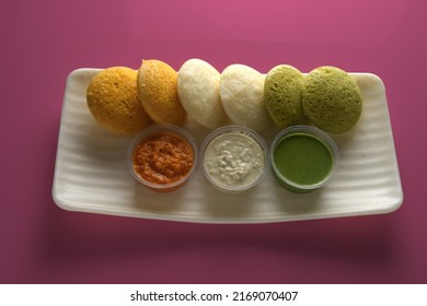 Tricolor Color Idli Blend Of Turmeric, Coriander Leaves, Oats, And Rice With Coconut, Tomato, And Coriander Chutneys On A Seamless Soft Pink Color Background.