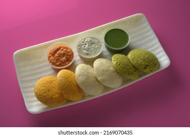 Tricolor Color Idli Blend Of Turmeric, Coriander Leaves, Oats, And Rice With Coconut, Tomato, And Coriander Chutneys On Seamless Soft Pink Color Background.