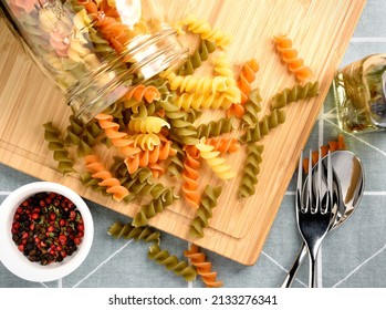 Tricolor dry pasta, rotini, fusilli pour from glass jar on wooden board with red and black pepper in white bowl and rosemary olive oil in glass bottle, top view, flat lay - Powered by Shutterstock