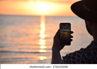 Trinidad, Cuba - January 2020: Silhouette Of A Man Wearing A Hat And Taking A Picture Of A Beautiful Sunset On The Beach. Sun Reflecting On The Water. IPhone Photography