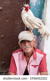 TRINIDAD, CUBA - MARCH 2, 2021: Portrait Of An Old Cuban Man With A Pet Chicken On His Head.