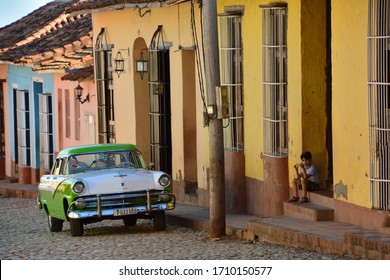 Trinidad, Cuba-March 2019: Street View Of Cobblestone Street With Classic Vintage Car In Front Of Colonial Houses With Little Kid Eating Sandwich On The Steps Of His House