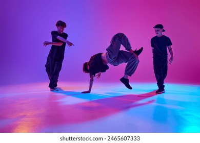 Trio of energetic teenagers, talented break-dancers dancing in motion in mixed neon light against vibrant gradient background. Concept of sport and hobby, music, fashion and art, movement. Ad