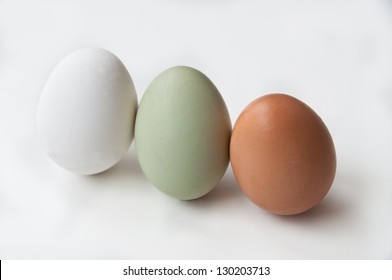 A trio of farm fresh eggs stands on a white background. In addition to the ordinary white egg is a green Araucana egg, and a brown Rhode Island Red egg.