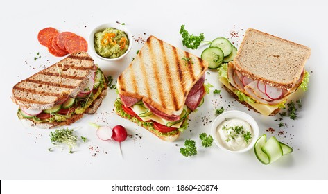 Trio Of Gourmet Sandwiches On Assorted Bread With Meat, Cheese, Guacamole And Pesto Fillings Surrounded By Ingredients And Seasoning On White In A Panorama Banner