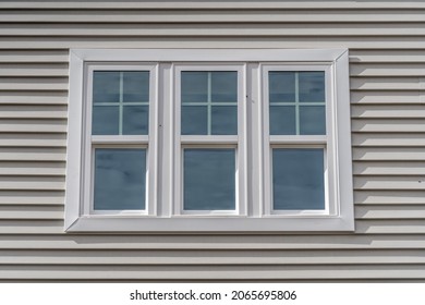 Triple hung window with fixed top sash and bottom sash that slides up, sash divided by white grilles a surrounded by white elegant frame  horizontal white vinyl siding on a new construction residence