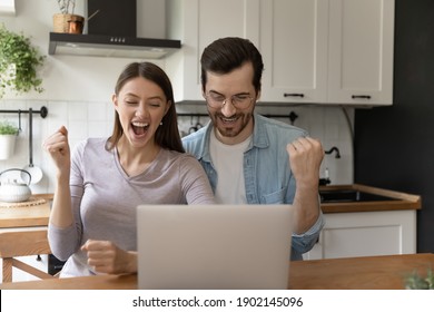 Triumph. Amazed Young Family Couple Sitting On Kitchen Screaming Of Joy By Laptop Screen Getting Good News. Excited Spouses Overwhelmed With Success Receiving Bank Credit On Family Business Developing