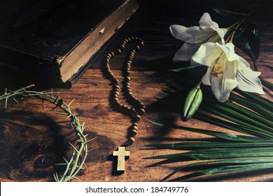 Triumph, Passion, Crucifixion And Resurrection Concept.  Iconic Symbols Relating To Palm Sunday And Easter Rest Upon A Rustic Table - Bible, Palm Branch, Crown Of Thorns, Cross, And White Lily.     