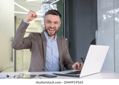 A triumphant businessman raises his fist in a gesture of success while working on his laptop in a modern office setting. - Powered by Shutterstock