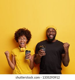 Triumphing glad ethnic couple clench fists, being in high spirit, listen favourite music, celebrate successful day, drink coffee or tea isolated over yellow wall with free space above. Leisure concept