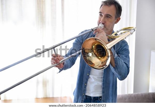 Trombonist practicing\
trombone in a room with curtain in the background at home. \
Horizontal\
composition.
